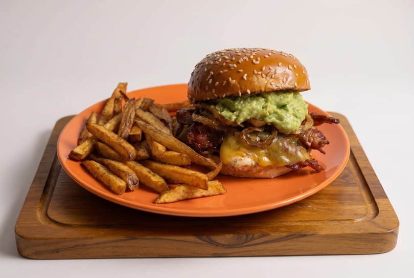 A gourmet burger with guacamole, caramelized onions, and bacon, served with fries on an orange plate.